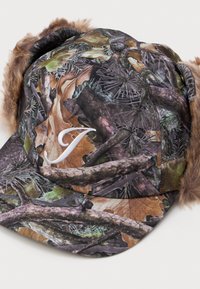 Klobúk so vzorom camouflage v hnedých a zelených odtieňoch, ktorý má umelecko-kožušinové uši a na prednej strane biely vyšívaný písmeno "J".