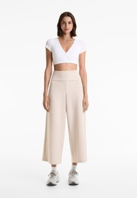 Witte, kortmouwige wikkeltop gecombineerd met hoog ingevoerde, beige ribgebreide wijde broek. Model draagt lichte sneakers en witte sokken.