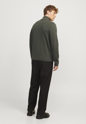 Donkergroen gebreide trui met hoge kraag, gedragen met zwarte broek en zwarte schoenen, gezien vanaf de achterkant. Glad oppervlak, relaxte pasvorm.