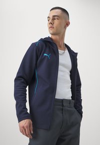 Navy zipsový mikina vyrobená z mäkkej, textúrovanej látky s svetlomodrými akcentmi a logom Puma. Obsahuje štandardný strih a bočné vrecká.