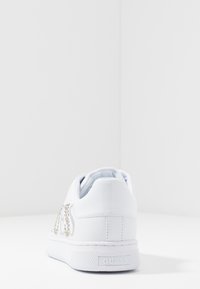 Baskets blanches en matière synthétique lisse, logo décoratif en strass, semelle blanche texturée et col rembourré pour le confort.