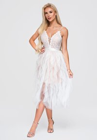 Femme blonde portant une robe blanche sans manches en dentelle et tulle avec des bretelles spaghetti et des sandales argentées à talons hauts, debout sur un fond uni.
