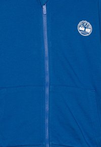 Sudadera con cremallera en color azul, con una textura suave, bolsillos tipo canguro y un logo de árbol blanco en el lado izquierdo del pecho. Se pueden ver la cremallera y las costuras.