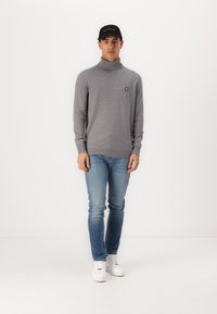 Grijze coltrui met een klein geel logo, gedragen met lichtblauwe jeans en witte sneakers. Het model draagt ook een zwarte pet.