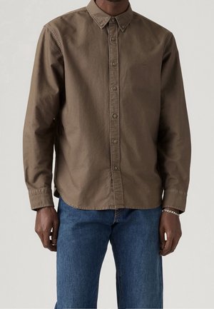 Man draagt een bruin overhemd met knopen en lange mouwen met een kraag, gecombineerd met een blauwe spijkerbroek en een zilveren armband om de rechterpols.