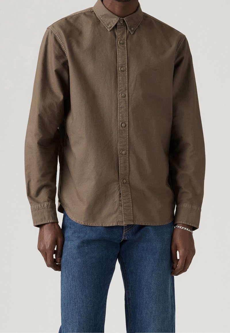 Homme portant une chemise marron à manches longues avec boutons et col, associée à un jean bleu et un bracelet argenté au poignet droit.