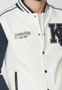 Veste universitaire blanche avec manches bleu marine, col côtelé. Présente une broderie "Legendary Since 2001" et une fermeture à boutons. Texture de tissu douce.