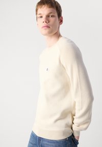 Ung mann med kort hår iført en kremfarget strikkegenser og blå jeans, stående mot en ensfarget lys bakgrunn.