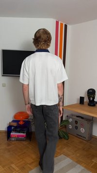 Biała, krótka, krótkorękawowa koszula o kroju typu crop top z kontrastującym granatowym kołnierzykiem, zestawiona z ciemnoszarymi dżinsowymi spodniami, widziana od tyłu.