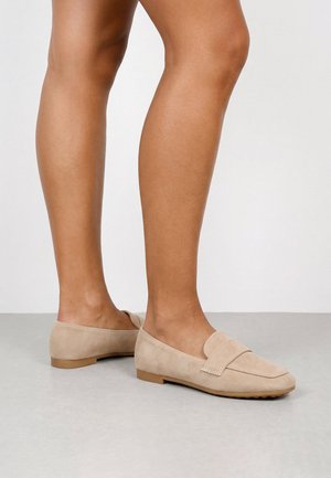 Mocasines de ante beige para mujer, usados con los pies descalzos, de pie sobre un suelo de color claro contra una pared blanca lisa.