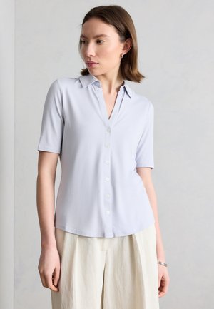 Woman with short brown hair wearing a light blue short-sleeve button-up shirt and cream wide-leg pants, looking to her left.