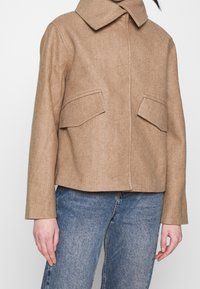 Brun tekstureret uldjakke med krave, forlommer og en kort pasform, parret med lyseblå jeans.