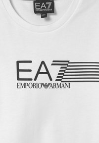 Vit skjorta med svart "EA7 Emporio Armani"-logotyp, inklusive stiliserad "7" med horisontella linjer och Emporio Armani-örnsymbol.
