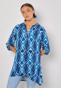 Blaue und weiße Tunika mit geometrischem Muster, V-Ausschnitt und hochrollbaren Ärmeln. Aus leichtem Stoff mit lockerer Passform gefertigt.