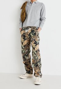 Mann trägt ein hellgraues Langarm-Polo, cargo-Hosen mit Camouflage-Blättermuster, weiße Sneaker und hat eine braune Jacke über die Schulter gelegt, vor einer weißen Wand.