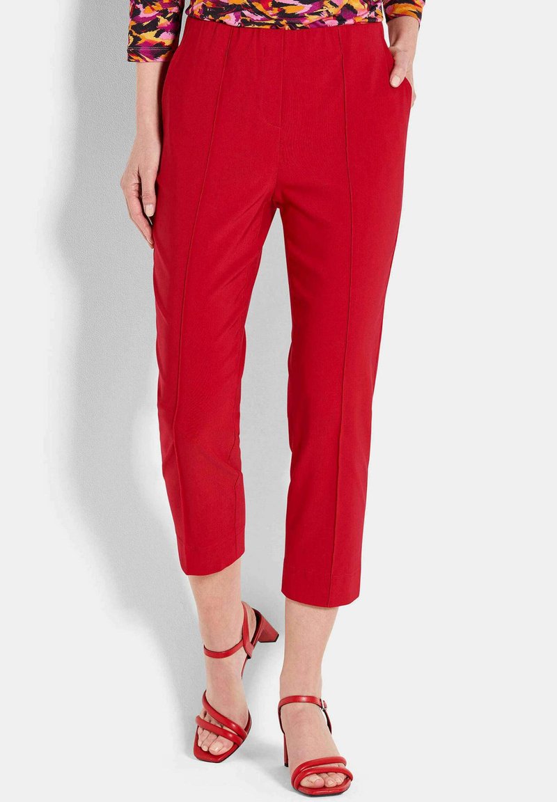 Frau in enganliegenden roten cropped Hosen mit vorderen Nähten, kombiniert mit roten Absatzsandalen und einem bunten gemusterten Oberteil.