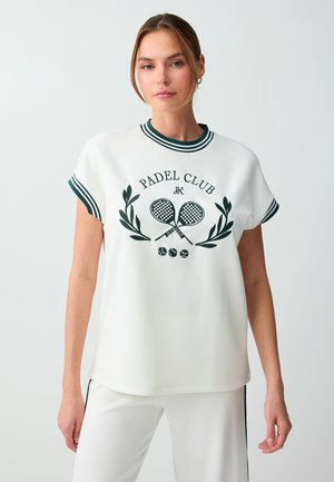 Jonge vrouw draagt een wit padelclub T-shirt met gestreepte kraag en mouwen, gecombineerd met een witte broek tegen een effen lichte achtergrond.