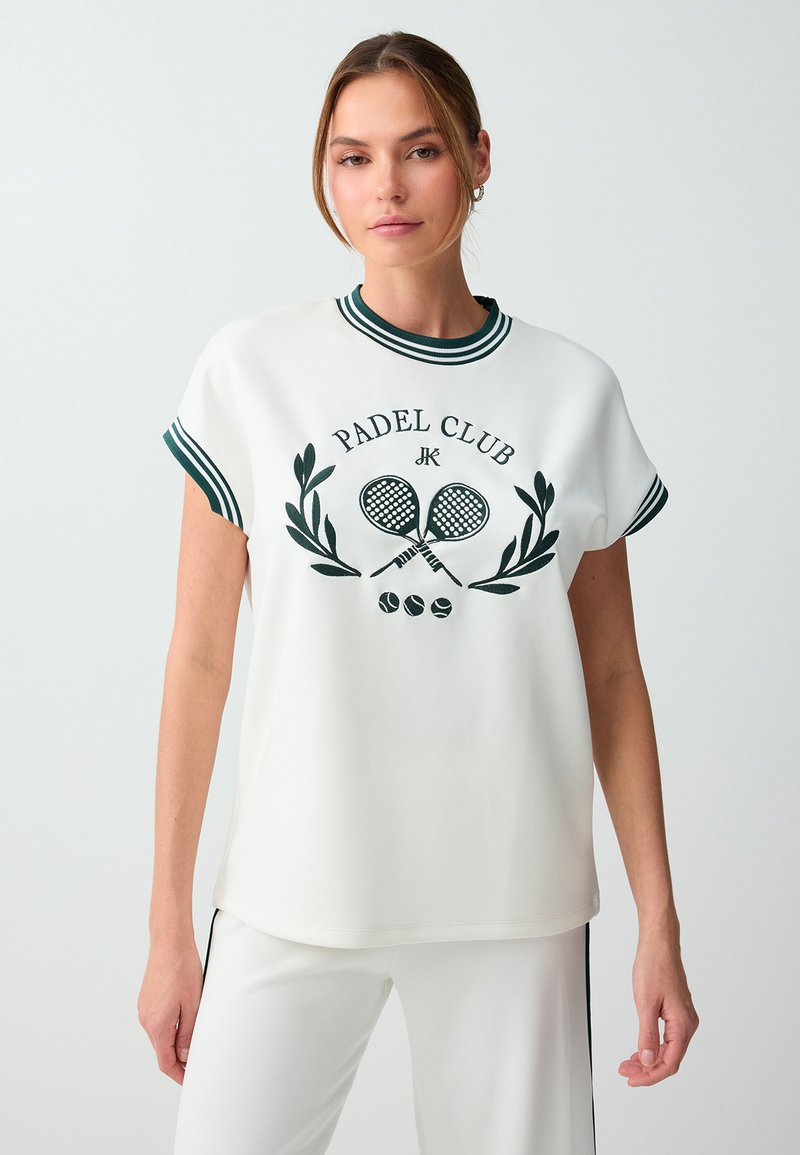 Jeune femme portant un t-shirt blanc du club de padel avec col et manches rayés, associé à un pantalon blanc sur un fond clair uni.