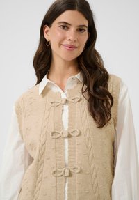 Ženska z dolgimi rjavimi lasmi, ki nosi zlate obročaste uhane, belo srajco s kratkimi rokavi in svetlo bež pleteno jopico z tremi sprednjimi pentljami.