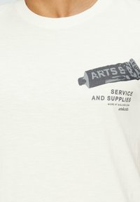 Witte katoenen t-shirt met een grijze gedrukte afbeelding van een verf tube met de tekst "KUNST & KLUSJES SERVICE EN BENODIGDHEDEN" eronder.