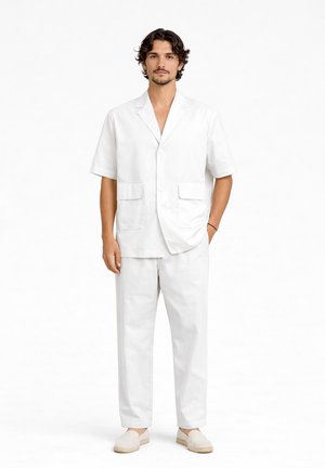 Uomo con capelli corti e ricci che indossa un blazer bianco a maniche corte, pantaloni abbinati e scarpe bianche slip-on, in piedi davanti a uno sfondo bianco.