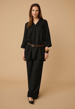 Donna con top nero scuro a V largo con cintura marrone, pantaloni ampi abbinati, scarpe nere a punta e bracciali spessi, in piedi su sfondo beige.
