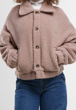 Nainen, jolla on beige nappikiinnitteinen sherpa-takki, kädet sivutaskuissa, yhdistetty tummansinisiin farkkuihin ja valkoiseen pooloneuleeseen alla.