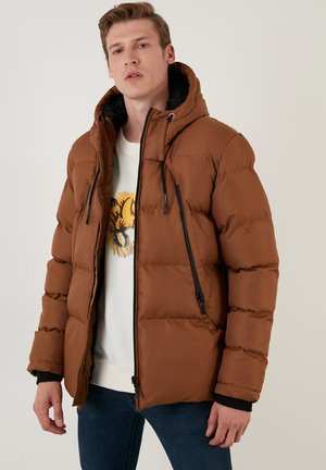 Brun pufferjakke med hætte, med lynlåslommer i siden, elastiske manchetter og en mat finish. Båret over en lysefarvet grafisk sweater.