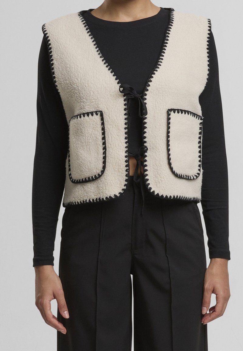 Personne portant un gilet en polaire crème avec des bordures de couture noires et des poches avant, par-dessus une chemise noire à manches longues et un pantalon noir.