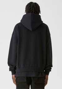 Melna kapuce, kas izgatavota no mīkstas auduma, ar plašu kapuci, brīvu piegriezumu, zemu plecu līniju un ribotām piedurknēm un apakšmali.