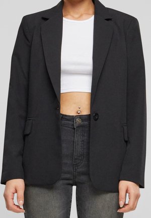 Personne portant un blazer noir, un crop top blanc laissant apparaître un piercing au nombril, et un jean taille haute gris foncé sur un fond uni.