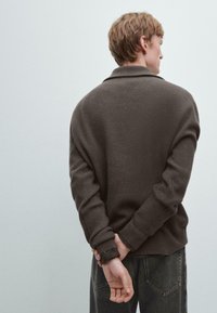 Braunes langärmeliges, geripptes Shirt mit lockerer Passform. Mit spitzem Kragen und umgeschlagenen Manschetten, kombiniert mit dunklen Denim-Jeans.