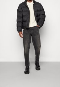 Černá prošívaná bunda s vysokým límcem, bílý pletený svetr, šedé džíny a černé pevné boty. Texturované materiály a moderní střih.