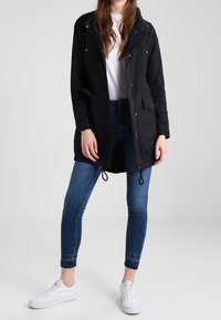 Černá lehká parka s předním zipem, stahovacími šňůrkami v pase, dvěma předními kapsami a kapucí, v kombinaci s modrými džíny a bílými teniskami.