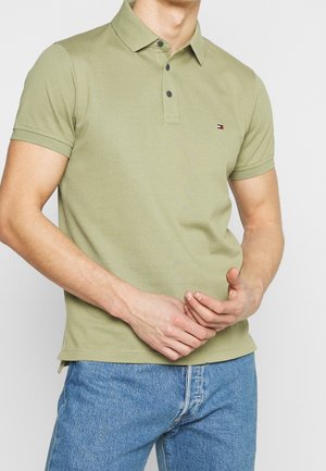 Mężczyzna ubrany w jasną oliwkowozieloną koszulkę polo z krótkim rękawem, z dwoma guzikami przy plisce i małym logo, zestawioną z niebieskimi jeansami.