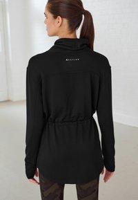 Haut de sport noir à manches longues avec un col haut, détail de taille cintrée et logo "ACTIVE" dans le dos, porté avec des leggings à motifs.
