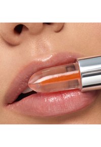Glossy lips apply transparent lipstick with an orange streak inside a silver tube, close-up on mouth and nose area.
