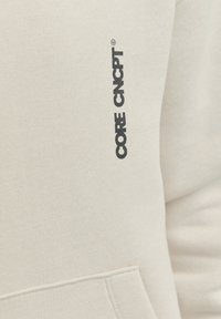 Bež dukserica s mekim teksturom, s crnim logotipom "CORE CNCP" na rukavu. Ima kengursku džep i rebraste rukave.
