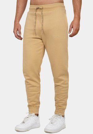 Mężczyzna ubrany w beżowe spodnie dresowe z troczkiem i białe trampki, stoi z rękami luźno opuszczonymi wzdłuż ciała na jednolitym tle.
