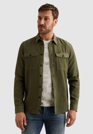 Mann trägt ein olivgrünes Hemd über einem gemusterten weißen T-Shirt und blauen Jeans, stehend vor einem hellgrauen Hintergrund.