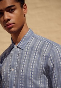 Chemise bleu clair à boutons avec un motif floral et des rayures verticales. Col court et coupe standard avec une texture douce.