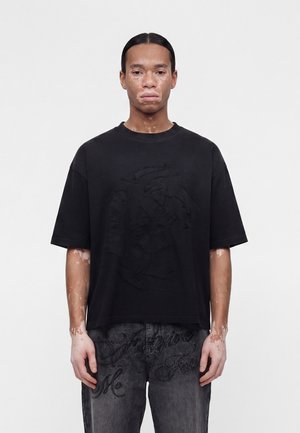 Giovane con vitiligine, che indossa una maglietta nera oversize e jeans scuri con ricami di lettere, in piedi contro uno sfondo chiaro e semplice.