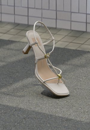 Sandale bej cu toc și barete, cu cataramă aurie și bilă decorativă aurie, pe pavaj lângă un perete din plăci.