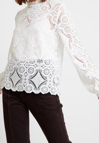 Femme portant un chemisier en dentelle blanche à manches longues avec des motifs floraux et géométriques, assorti à un pantalon moulant foncé.