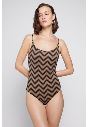 Žena s krátkými hnědými vlasy, nosící zlaté kruhové náušnice a hnědočerný bezrukávový overal se vzorem cikcak, s rukama za zády.