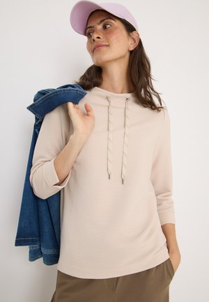 Beige Kapuzenoberteil mit Rippstruktur, Kordeln mit metallischen Enden. Kombiniert mit einer Denimjacke über der Schulter und neutralfarbenen Hosen.