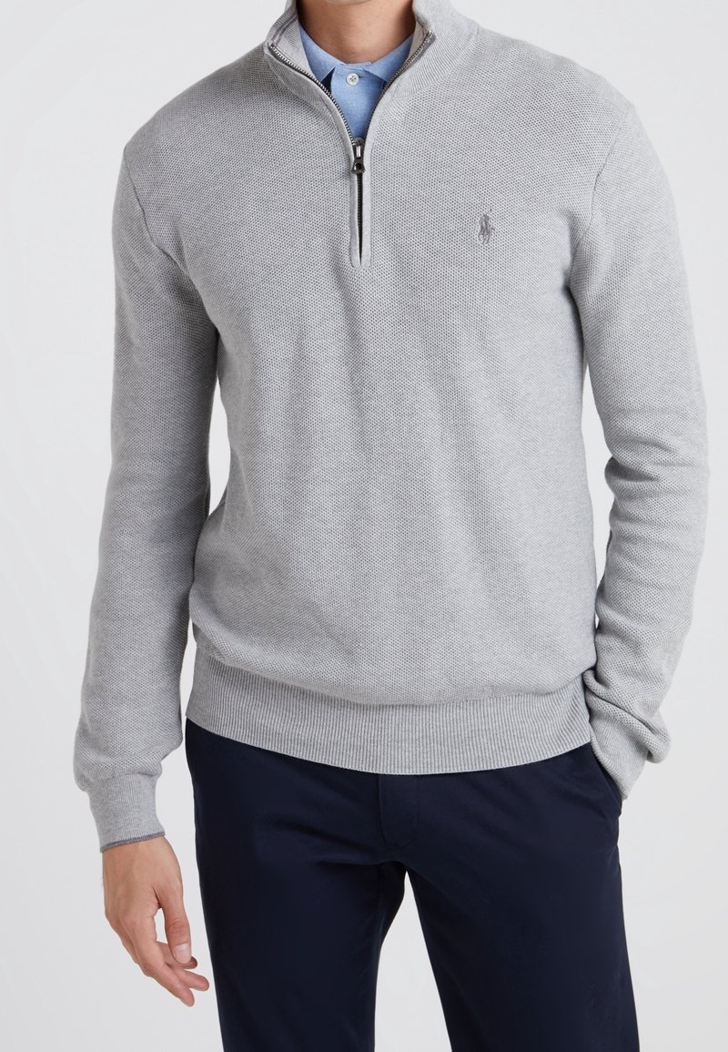 Grå zip-up tröja med texturerad yta, ribbade ärmslut och nederkant, med en liten logotyp på bröstet, bärs över en ljusblå skjorta.