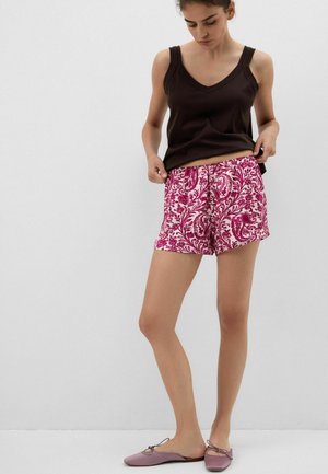 Kobieta poprawiająca różowo-białe krótkie spodenki w kwiatowy wzór, ubrana w czarny top bez rękawów i różowe płaskie buty, stoi na tle jednolitego szarego tła.