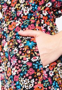 Tela con estampado floral en negro con flores multicolores. Una mano se asoma a un bolsillo lateral, mostrando el diseño del bolsillo.