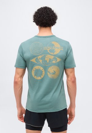 Άνδρας με σγουρά μαλλιά που φοράει τιρκουάζ μπλουζάκι με ξεθωριασμένο κίτρινο σχέδιο παγκόσμιου χάρτη, ήλιου, φεγγαριού και κυκλικών σχεδίων στην πλάτη, σε συνδυασμό με μαύρα σορτς.